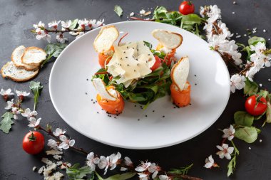 Beyaz tabakta yeşil sarılmış ve peynirle kaplanmış somonlu bir salata var. Tabağı çevreleyen kiraz domatesleri ve otlar, tabakların koyu bir masa üzerindeki görüntüsünü güçlendiriyor..