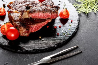 Orta pişmiş bir biftek taze vişneli domateslerle çevrili siyah bir tahtada duruyor. Yakınlarda bir çatal var. Bu sahne mutfağın tadını çıkarmaya hazır bir yemeği yakalar..