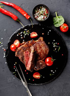 Izgara biftek, içinde kiraz domatesleri, taze kırmızı biber ve küçük bir kase baharat olan siyah bir tabakta düzenlenir. Dekorasyonda zevke hazır lezzetli bir yemek sergileniyor.