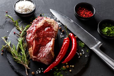 Bifteğin taze bir kesimi, otlar ve kırmızı biberlerle çevrili koyu bir tahta yüzeyde oturur. Keskin bir bıçak ve küçük bir kase tuz ve biber de mevcut..