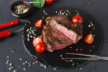 Pişmiş bir biftek kesilir ve siyah bir taş tabağa yerleştirilir. Bifteğin etrafı kiraz domatesleriyle çevrili. Arka planda küçük bir tas baharat var. Bu sahne bir mutfakta geçiyor..