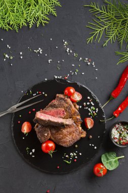 Koyu bir tabakta bir biftek, kiraz domatesleri ve taze otlarla servis edilir. Biftek sulu pembe bir merkez gösteriyor. Etrafında baharatlar ve acı biberler var..