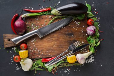 Tahta kesme tahtasında çeşitli taze sebzeler sergileniyor. Sebzelerin yanında bir çatal ve bıçak var. Mutfak ortamında yemek pişirme hazırlığı öneriliyor..