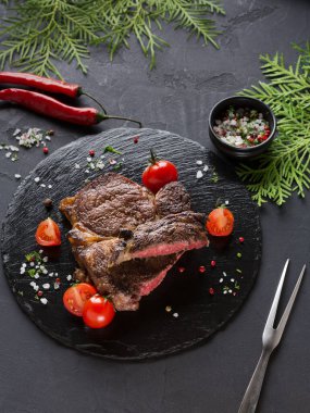 Izgara biftek, kiraz domatesleri ve yeşil otlarla çevrili siyah bir tabağa konur. Kırmızı biber ve bir kase tuz yakında. Yemek için hazır ve iştah açıcı görünüyor..