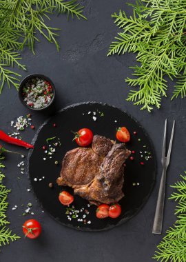 Izgara bir et parçası siyah bir taş tabağa konur. Etin etrafı kiraz domatesleriyle çevrili ve yanında baharatlı küçük bir kase var. Yeşil yapraklar sahneyi çerçeveler.