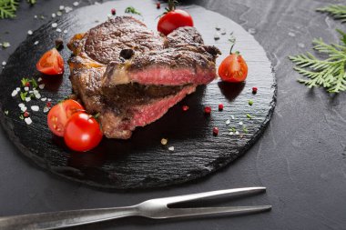 Izgara bir biftek siyah bir taş tabakta oturur. Etin etrafı kiraz domatesleri ve yeşil otlarla çevrili. Çatallar yakınlarda. Keyfini çıkarmaya hazır bir yemeğe benziyor..