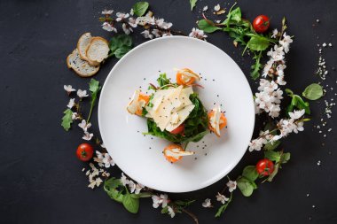 Bir tabakta yeşillik, domates, peynir ve otlardan oluşan bir salata vardır. Tabağın etrafında ekmek parçaları ve çiçek yaprakları var. Bu karanlık bir yüzeye ayarlanmış..