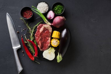 Çeşitli taze malzemeler yemek pişirmeye hazır siyah bir masaya konur. Bunlar et, sebze, bitki ve baharattır. Kesmek için yakına bir bıçak konur..