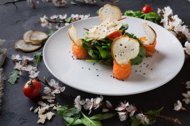 Salata yeşillikleri, somon dilimleri ve patates kızartması şeklinde kızarmış ekmek içeren bir tabak beyaz bir tabakta düzenlenir. Çeri domatesler ve çiçekler koyu bir yüzeyde tabağın etrafını sarar..