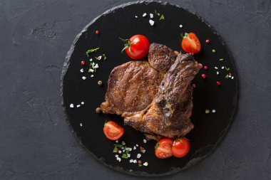 Izgara biftek siyah taştan bir tabağa konur. Bifteğin etrafı kiraz domatesleriyle çevrili. Sahnede yemeğin etrafına saçılmış tuz, bitki ve renkli biber var..