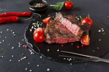 Dilimlenmiş ızgara biftek. Bifteğin etrafına taze domates ve kırmızı biber dizildi. Siyah taş tabağı karanlık bir yüzeye yerleştirildi. Yakınlarda bir kase baharat var..