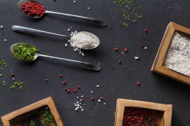 Karanlık bir yüzeye yerleştirilmiş farklı tuz ve baharat türleri. Tuz ve biber içeren ahşap kaplar ve etrafa saçılmış bazı otlar var. Kaşıklar malzemelerin karışımını barındırır..