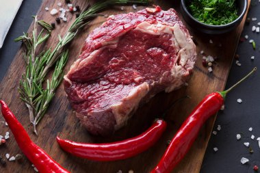 Biberiye ve parlak kırmızı biber filizleriyle çevrili ahşap bir tahtanın üzerinde taze bir et parçası duruyor. Sahne, yemek pişirmeden önce hazırlanmış malzemeleri yakalar..