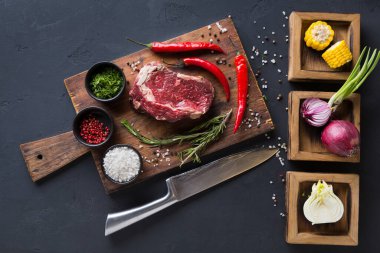 Bir parça çiğ et, etrafını baharatlar, otlar ve sebzelerle dolu bir kesme tahtasına konur. Sahne yemek hazırlığı için hazır bir mutfak düzenini gösteriyor..