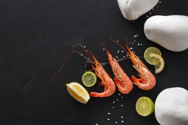 Limon dilimleri ve deniz tuzu ile birlikte siyah bir yüzeye üç karides yerleştirilir. Arka planda beyaz taşlar görülüyor, sahneye basit bir karşıtlık ekleniyor.