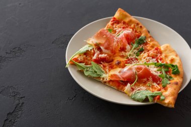 Bir dilim pizza sade beyaz bir tabakta oturur. Pizzada jambon, roka ve peynir gibi malzemeler var. Tabak karanlık bir yüzeyde duruyor, basit bir sunum oluşturuyor..