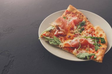 Bir dilim pizza beyaz bir tabağa konur. Pizzada jambon, peynir ve yeşillik var. Tabak karanlık bir yüzeyde duruyor..