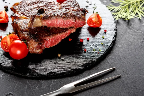 Orta pişmiş bir biftek taze vişneli domateslerle çevrili siyah bir tahtada duruyor. Yakınlarda bir çatal var. Bu sahne mutfağın tadını çıkarmaya hazır bir yemeği yakalar..
