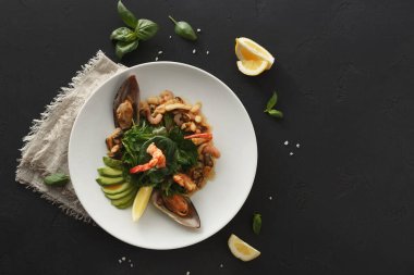 Bir deniz ürünleri tabağında servis edilir. Yemek karides, istiridye ve çeşitli sebzeleri içerir. Tabağın etrafına limon ve yeşil bitkiler konur..