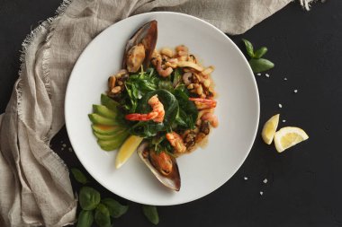 Bir tabakta karides ve midye içeren deniz ürünleri karışımı bulunur. Taze yeşil sebzeler deniz ürünlerinin etrafına dizilir. Limon ve otlar tabağın yanına konur..