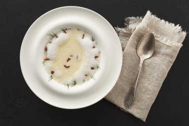 Bir kase, içinde bitkiler ve küçük kurutulmuş malzemeler bulunan temiz bir çorba tutar. Koyu bir masada katlanmış keten peçetenin yanında bir kaşık uzanıyor, yemek sahnesi yaratıyor..