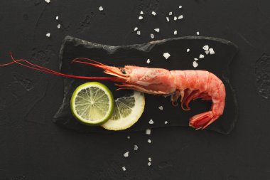 Büyük bir karides siyah bir taş levhanın üzerine yerleştirilir. Limon ve limon dilimleri karidesin yanında düzenlenir. Deniz tuzu kristalleri ekrana saçılmış..