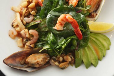 Deniz ürünleri tabağında karides, midye ve yeşil salata bulunur. Yemeğe bir dilim avokado ve bir dilim limon eşlik ediyor. Burası bir yemek salonu gibi görünüyor..