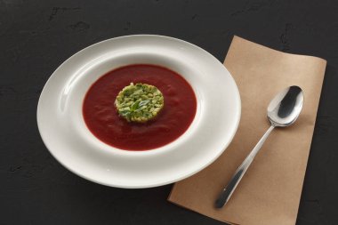 Beyaz bir kase, ortasında yeşil bir garnitür olan koyu kırmızı bir çorba tutar. Kase siyah bir masanın üzerinde, yanında basit bir kaşık ve kahverengi bir peçete ile duruyor..