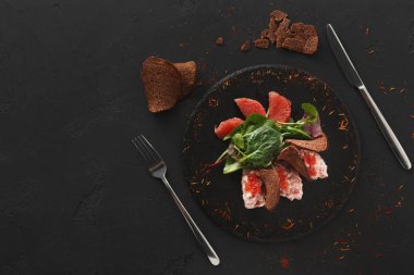 Bir tabakta karışık yeşillik, greyfurt segmenti ve çavdar ekmeğinden yapılmış taze bir salata var. Koyu bir masada tabağın yanına iki kap yerleştirildi..