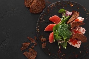 Yapraklı yeşillikli bir salata, bir dilim greyfurt ve bir parça kahverengi ekmek koyu servis tabağında sunulur. Yemekler basit bir şekilde düzenlenir..