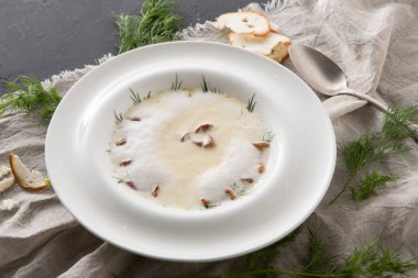 Bir kase kremalı çorba yakınlarda bir parça ekmekle kumaşın üzerinde duruyor. Çorbanın üstüne serpiştirilmiş otlar var. Bir kaşık kasenin yanında yatıyor, servise hazır..