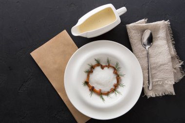 Koyu bir yüzeye krem bazlı bir tabak yerleştirilir. Sos yanında servis teknesinde duruyor. Taze otlar tabağın etrafında daire şeklinde dizilir. Yakınlarda bir kaşık duruyor..