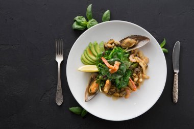 Bir deniz ürünleri tabağı karides, midye ve yeşillik içeren beyaz bir tabağa yerleştirilir. Tabak avokado dilimleri ve limon dilimleriyle süslenmiş. Yakınlarda çatal ve bıçak var..