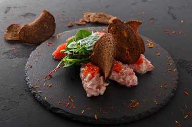 Koyu taştan bir tabakta yiyecek karışımı. İçinde et, kırmızı havyar, yeşil yapraklar ve kızarmış ekmek var. Ayarlar temiz ve basit görünüyor..