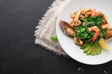 Deniz ürünleri salatası beyaz bir tabağa konur. Tabakta karides, midye ve taze yeşillikler yer alıyor. Avokado ve limon dilimleri salatanın etrafına dizilir..