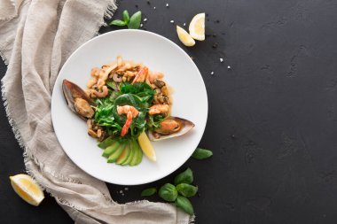 Bir tabakta karides, yeşillik, dilimlenmiş avokado ve limon içeren bir deniz ürünleri tabağı bulunur. Yemek sadece karanlık bir yüzeyde açık bir kumaş ile düzenlenir ve net bir sunum oluşturur..