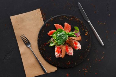 Tabakta balık, yapraklı yeşiller ve bir dilim meyve içeren bir salata var. Çatal bıçak takımı yakınlarda duruyor ve sete kahverengi kağıt peçete ekleniyor..