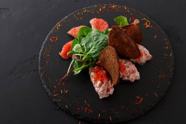 Siyah bir tabakta renkli bir tabakta kırmızı greyfurt, yapraklı yeşiller ve kahverengi ekmek bulunur. Tabakta çeşitli dokular ve tatlar var. Yaratıcı bir mutfak sunumu sergiliyor..