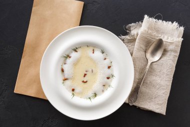 Bir kase çorba karanlık bir yüzeye konur. Çorbada hafif bir renk ve küçük bitkiler var. Bir kaşık kasenin yanında duruyor. Yakınlarda keten peçete ve kese kağıdı var..