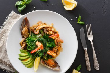 Bir tabakta karides, midye ve yeşillik içeren bir deniz ürünleri tabağı vardır. Yanında limon ve avokado dilimleri var. Çatal bıçak takımı tabağın yanına, karanlık bir masanın üzerine konur..