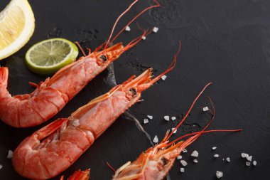 Taze karides, limon ve limon dilimlerinin yanına koyu bir yüzeye yerleştirilir. Deniz tuzu etrafa serpilmiş. Bu sahne deniz ürünleri hazırlamak ya da yemek tariflerini paylaşmak için idealdir..