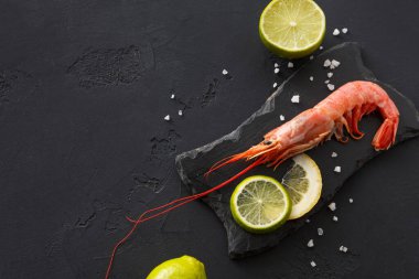 Dilimlenmiş limon ve limonla siyah bir levhaya karides konur. Kaba tuz deniz ürünlerinin etrafına serpilmiş, basit bir yiyecek düzenlemesi sergiliyor..