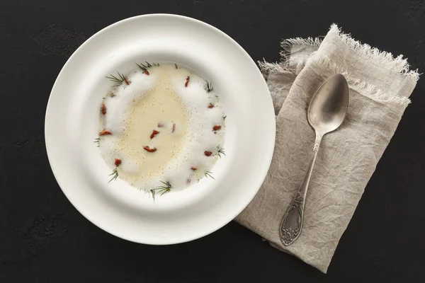 Bir kase, içinde bitkiler ve küçük kurutulmuş malzemeler bulunan temiz bir çorba tutar. Koyu bir masada katlanmış keten peçetenin yanında bir kaşık uzanıyor, yemek sahnesi yaratıyor..