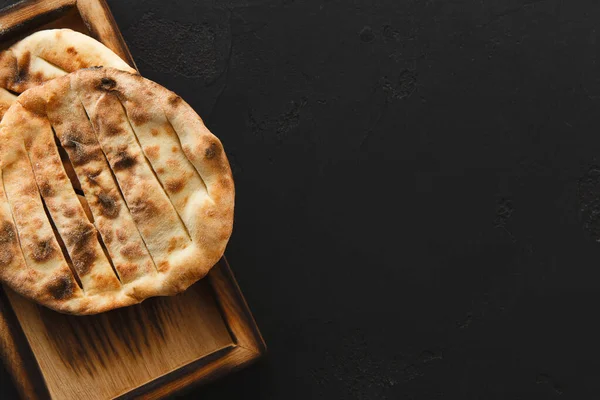 Tahta tepside oturan iki yuvarlak düz ekmek. Düz ekmekler ızgara izleri ve altın-kahverengi bir renk gösteriyor. Tepsi koyu bir yüzeyde duruyor, ekmeğin dokusunu ve şeklini vurguluyor..