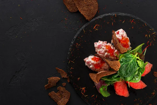 Bir tabak siyah ekmek dilimleri, kırmızı soslu bir karışım ve siyah tabakta taze yeşillikler içerir. Restoran ortamında servis ediliyor. Küçük ekmek parçaları etrafa saçıldı..