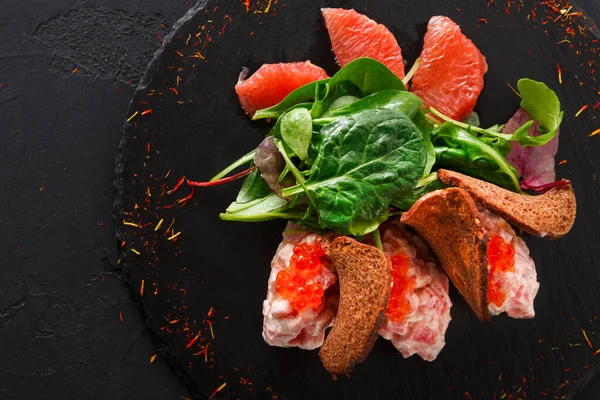 Tabakta taze ıspanak, greyfurt dilimleri ve siyah bir sayfada ekmekle sebze karışımı yer alıyor. Yemek hazırlandı ve bir yemek düzeninde servis edildi..