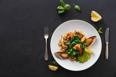 Tabakta taze sebzelerle çevrili karides ve midye karışımı yer alır. Limon dilimleri kenara konur. Masanın üzerinde bir çatal ve bıçak var..