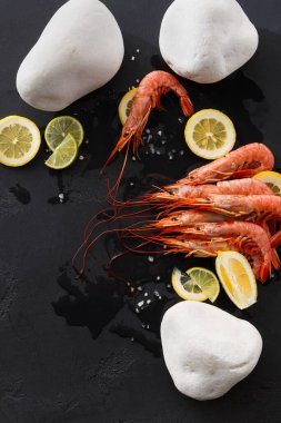 Karides, limon ve limon dilimlerinin yanına siyah bir yüzeye yerleştirilir. Beyaz taşlar deniz ürünlerinin etrafına dizilmiştir. Bu ayar yemek pişirmeye ya da yemek sunumuna odaklanmamızı öneriyor.