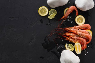 Karides dilimlenmiş limon ve deniz tuzu ile birlikte karanlık bir yüzeye yerleştirilir. Düzenleme beyaz taşları içeriyor, basit bir mutfak görüntüsü yaratıyor. Pişmeye hazır taze deniz ürünlerini gösteriyor..