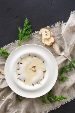 Bir kase çorba nötr keten kumaşın üzerinde duruyor. Çorbada bitkiler var ve yanında da bir dilim ekmek var. Yakınlarda bir metal kaşık duruyor, basit ve davetkar bir yemek takımı oluşturuyor..